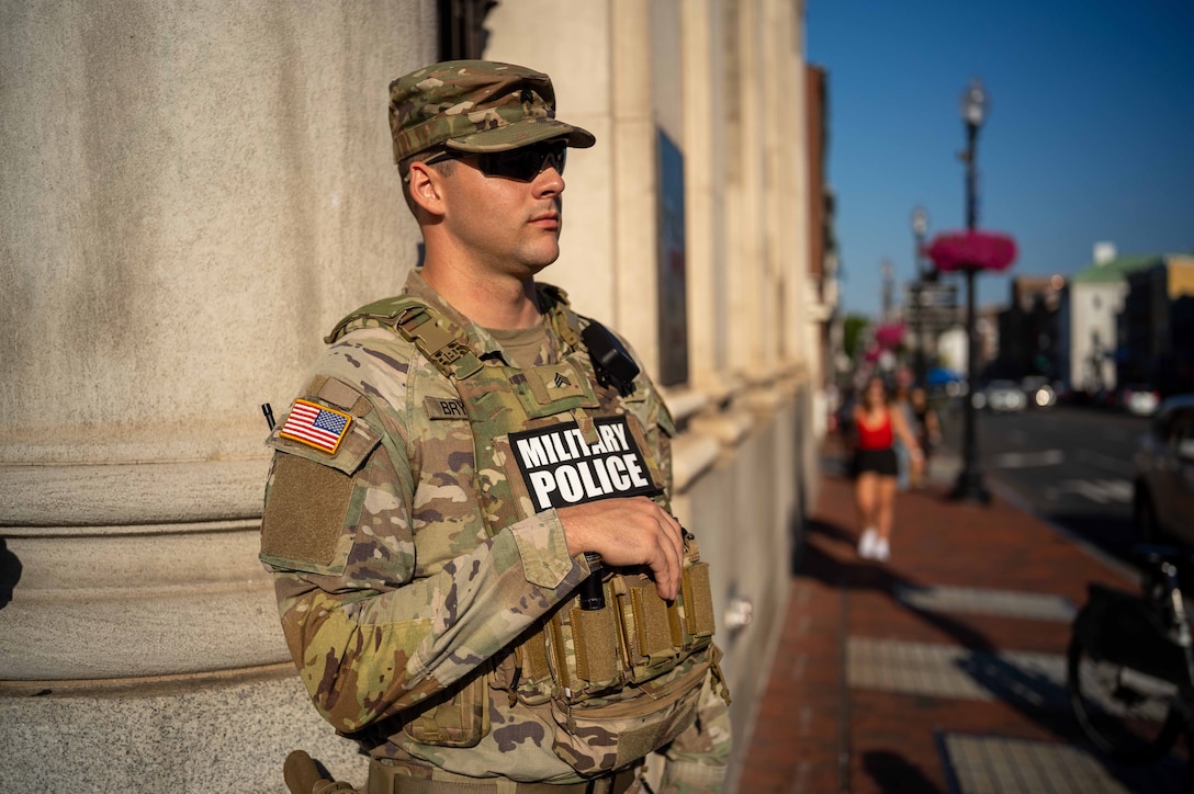 A person wearing a military camouflage uniform stands on a brick sidewalk in front of a building. People walk along the sidewalk in the background.