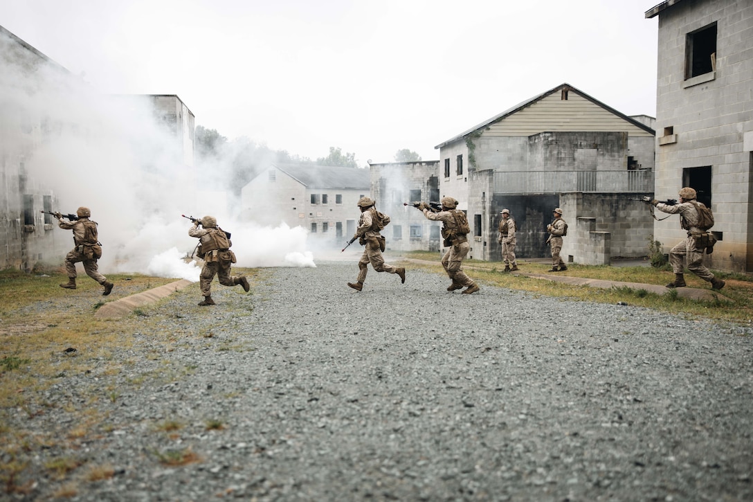 A group of Marines in tactical gear aim weapons as they run to the left across a gravel street with buildings lining both sides.