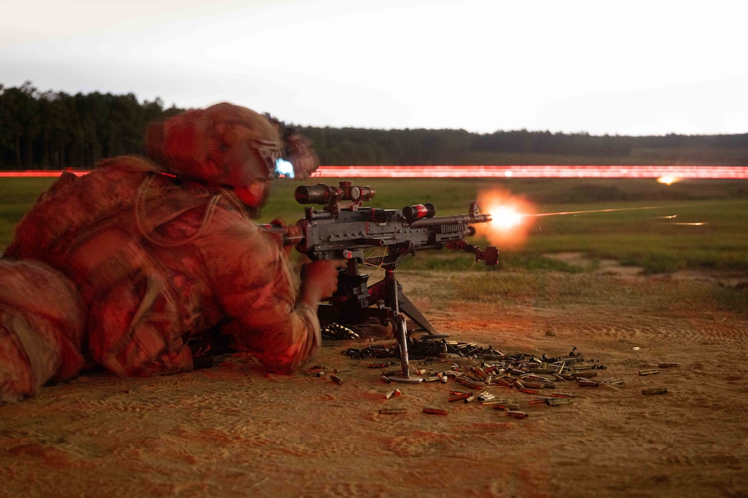 A soldier in tactical gear lays on a field and shoots a machine gun to the right, which leaves bursts and trails of red light.