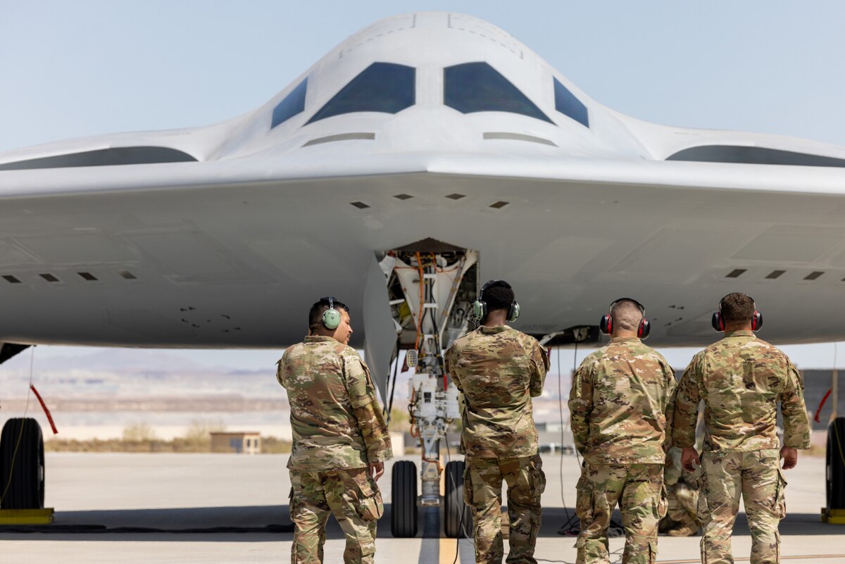 Four men wearing military camouflage uniforms and hearing protection stand in front of a large military bomber aircraft on a tarmac.