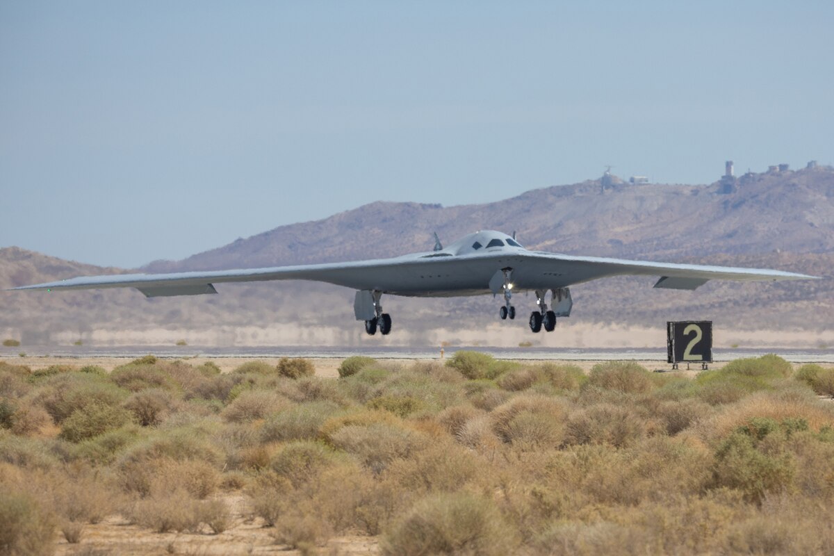 A military bomber aircraft with its wheels down prepares to land on a runway. There are mountains in the background and desert plants in the foreground.