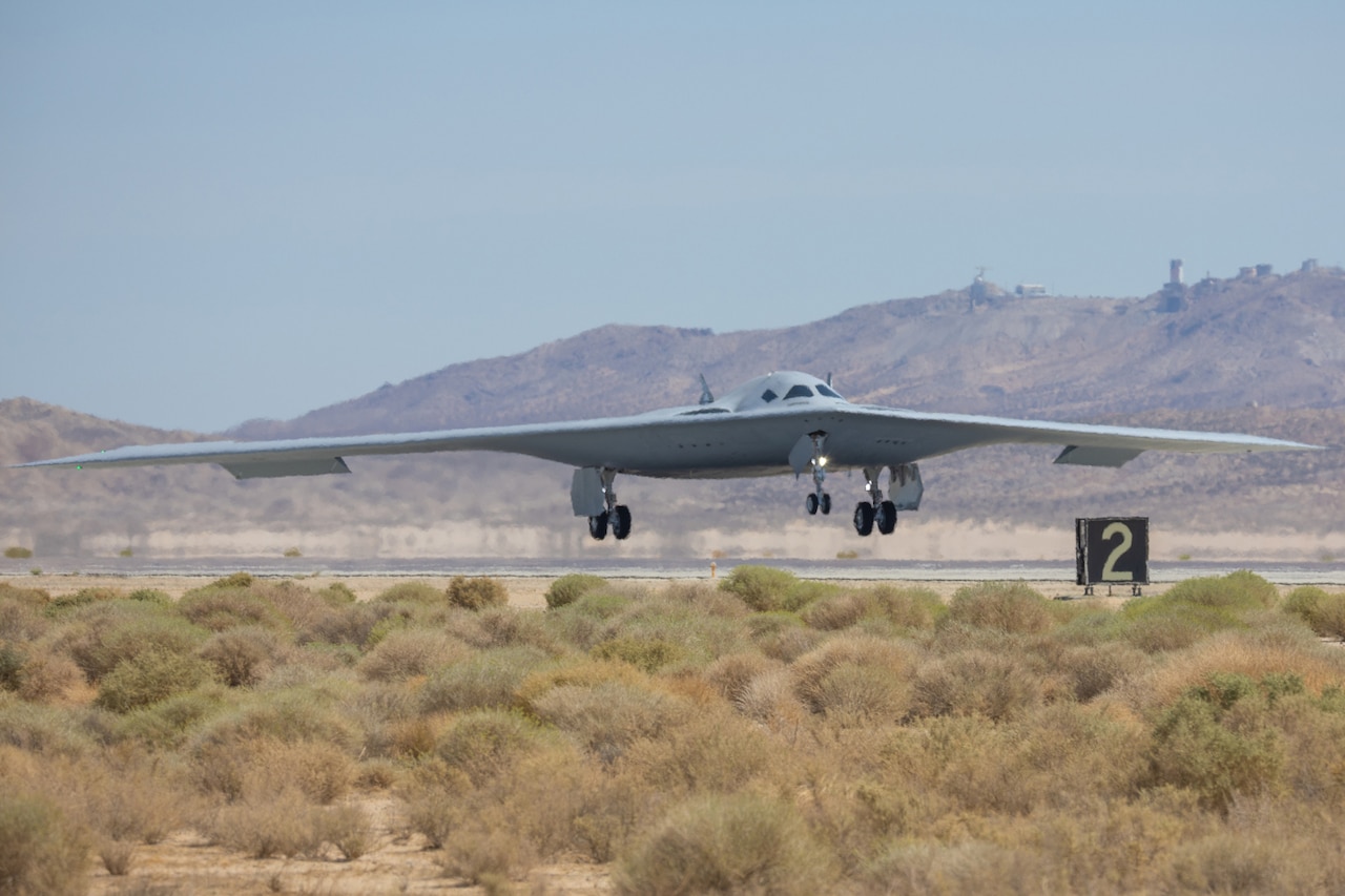A military bomber aircraft with its wheels down prepares to land on a runway. There are mountains in the background and desert plants in the foreground.