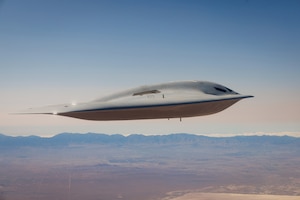 A military bomber aircraft flies over California. There is flat ground below the aircraft and mountains in the background.