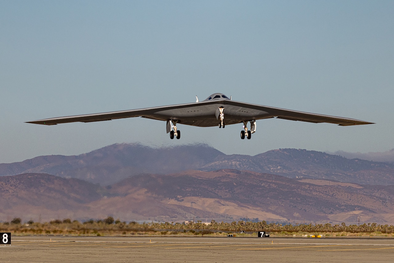 A military bomber aircraft with its wheels down prepares to land on a runway. There are mountains in the background.