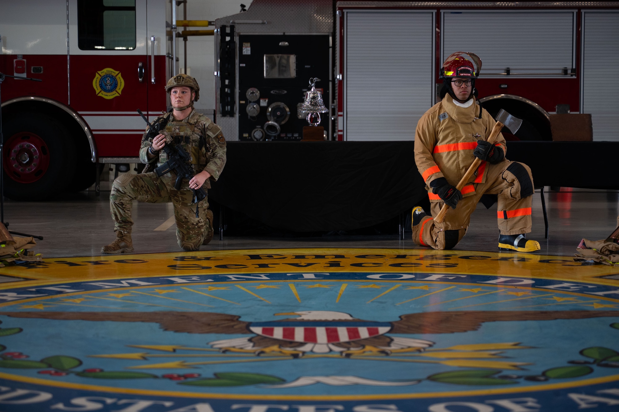 Two service members are kneeling down.  One is holding a weapon in their hand and the other, dressed in firefighting gear is holding an axe.  