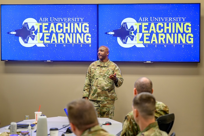 A Servicemember is standing in front of two screens briefing a crowd.