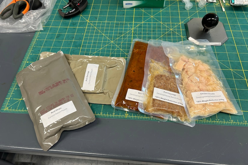 Four plastic packages sit on a table; Three are clear and show food contents inside.