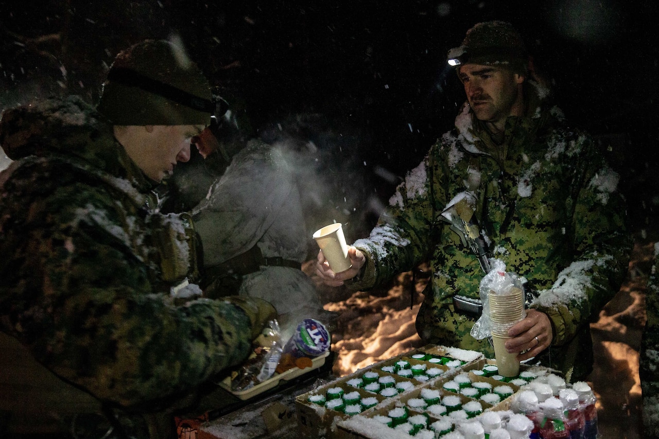 A man in a jacket covered in snow hands a tray with a cup to another man in a meal line.