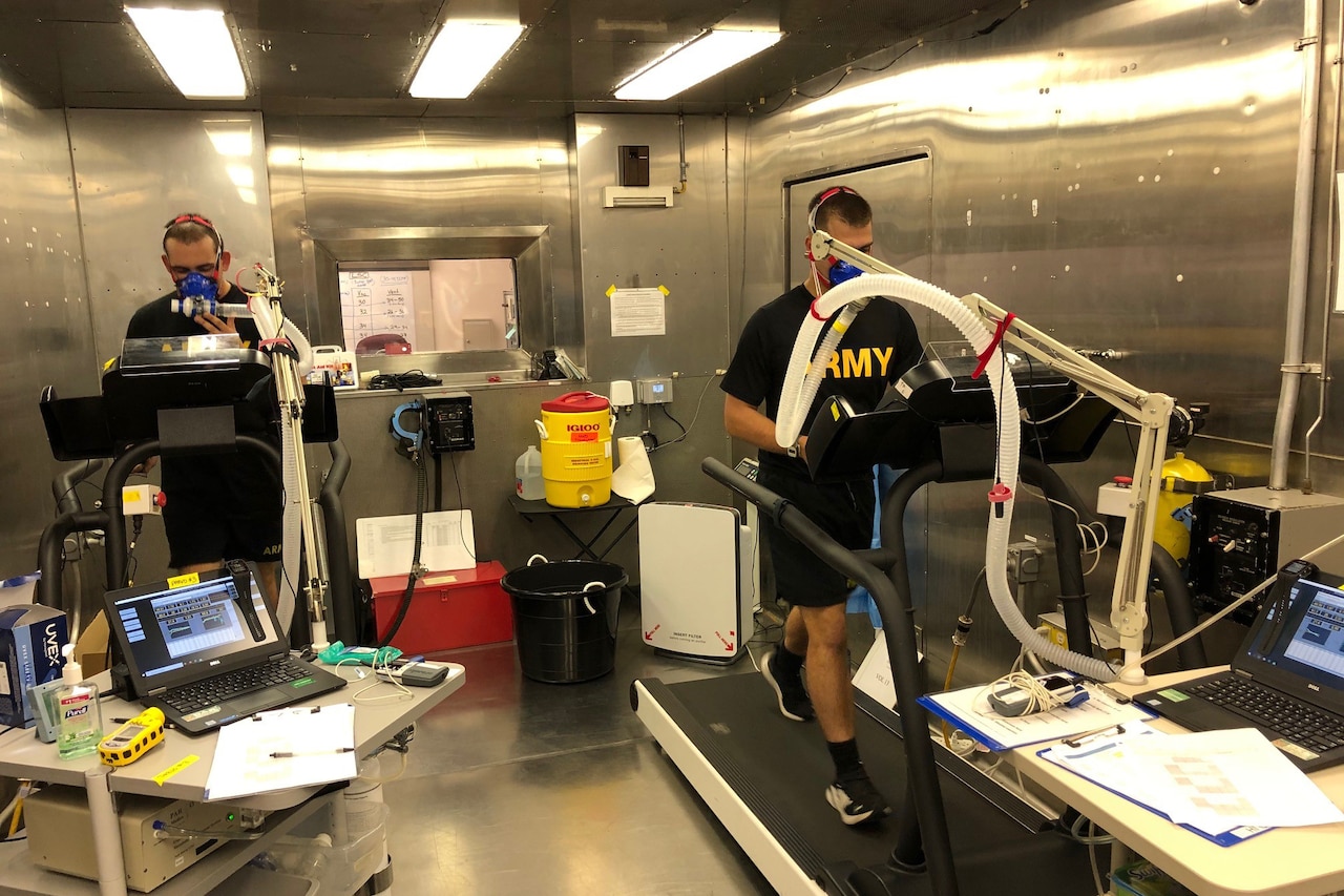 Two men wearing breathing apparatuses walk on treadmills in a small, metal room. Computer desks sit nearby.