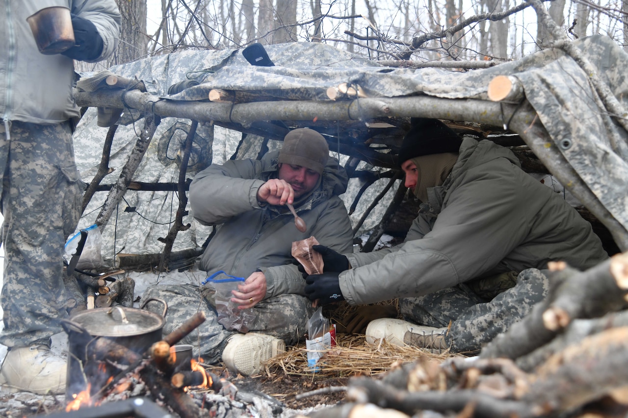 Two men share a small, packaged ration while huddled under a shelter made of logs and camouflage.