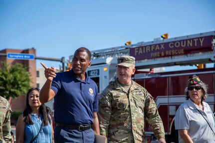 A man in a blue polo shirt is pointing to the right while walking with a U.S. Army Soldier who is wearing combat uniform during an outdoor event near a firetruck.