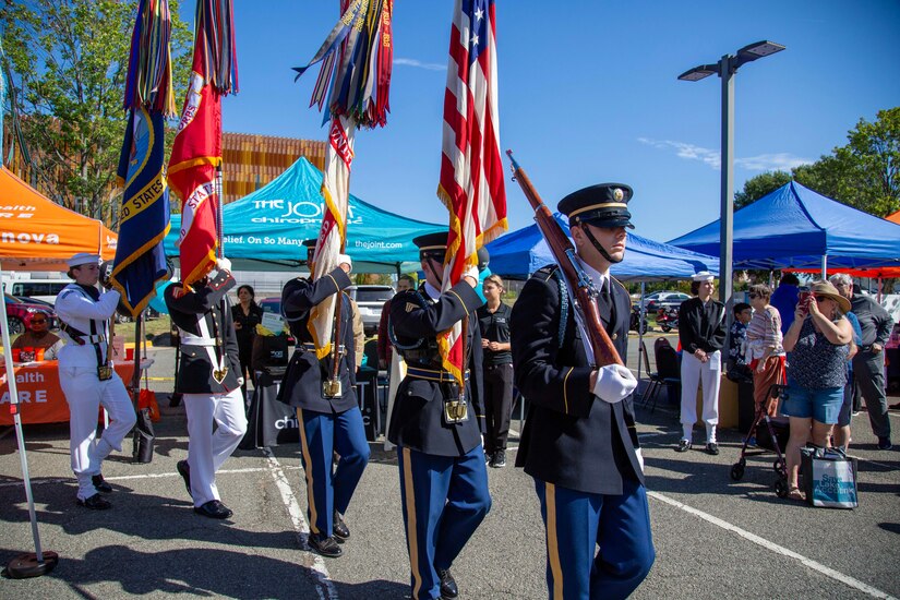 Military members carrying flags and rifles are marching during an outdoor ceremony.