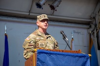Army Soldier wearing combat uniform is speaking at a wooden podium during an outdoor event.
