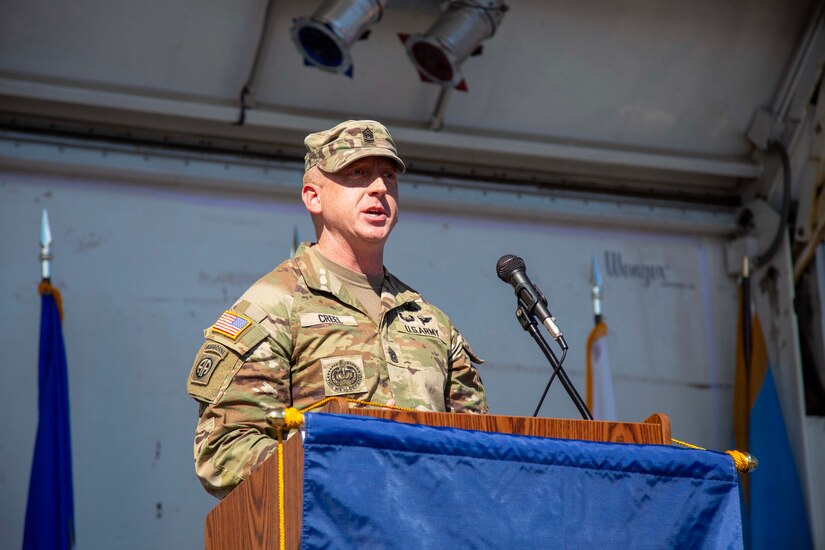 Army Soldier wearing combat uniform is speaking at a wooden podium during an outdoor event.