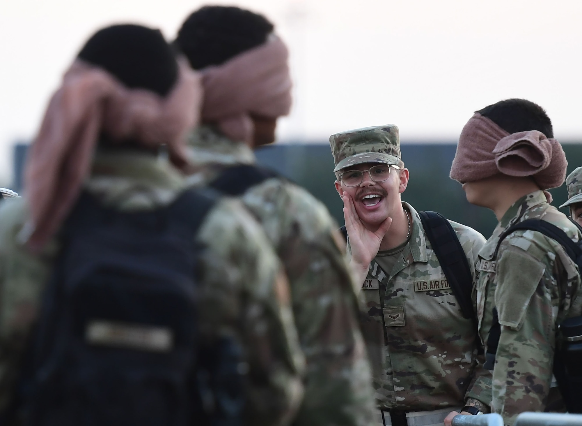 A man in military uniform shouts toward other men who are wearing towels over their eyes.