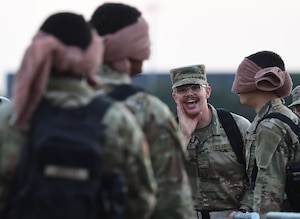 A man in military uniform shouts toward other men who are wearing towels over their eyes.