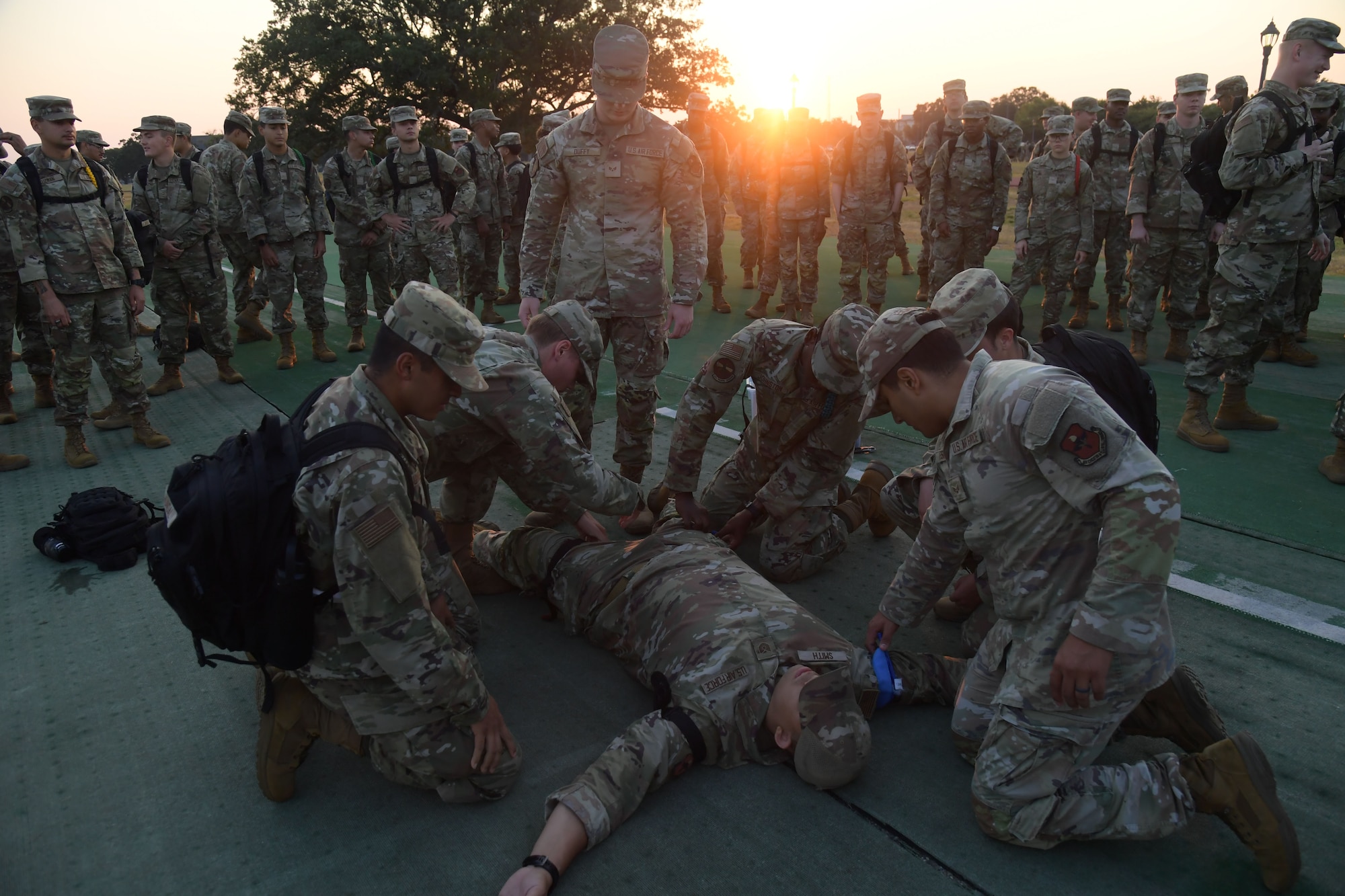A group of people in military uniform administer medical care to another in front of a larger group of people.