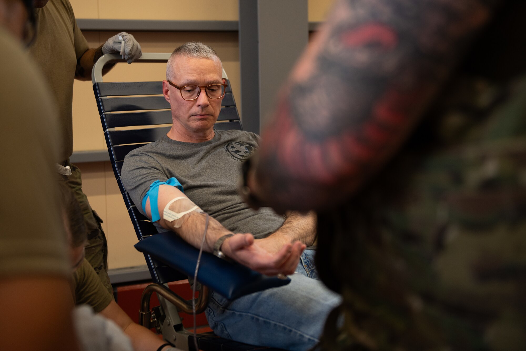 An Airman sits in a chair looking at their arm as they give blood during a blood transfusion.