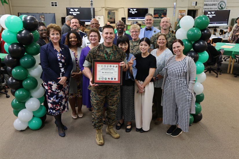 A man dressed in a military camouflage uniforms holds a certificate and is surrounded by people dressed in civilian attire and military camouflage uniforms. There is a balloon arch to the left and right of the group.