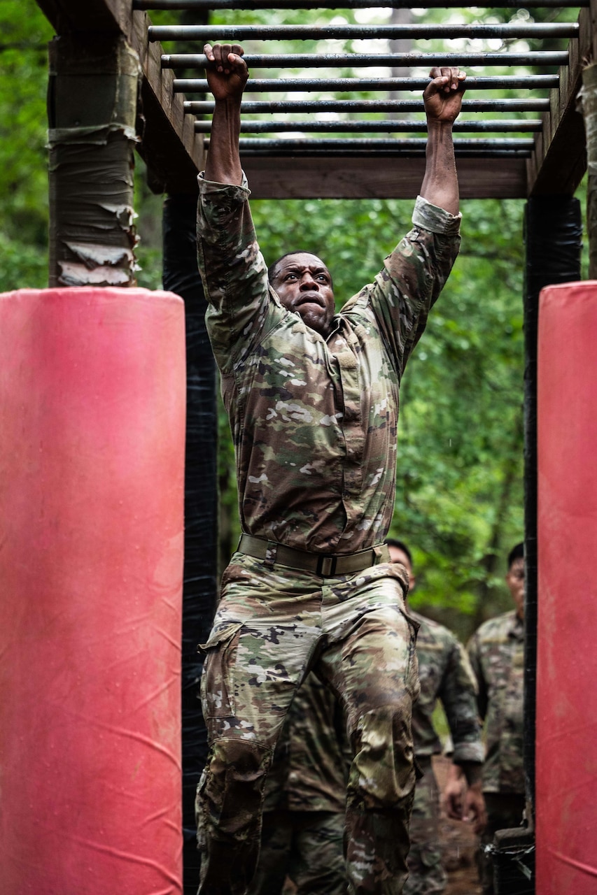 A person wearing a camouflage military uniform hangs from monkey bars as people in similar attire wait their turn on the obstacle.