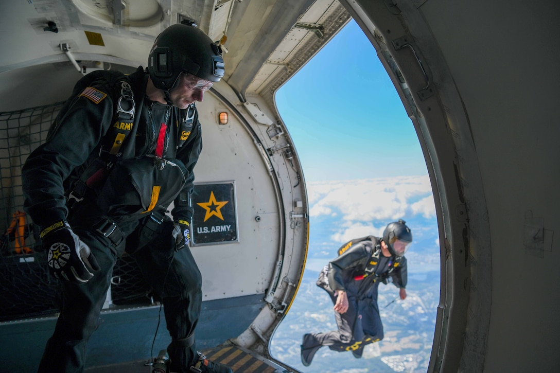 An Army parachute team member free-falls outside of an aircraft in a cloudy sky as a fellow team member watches from the open doorway.