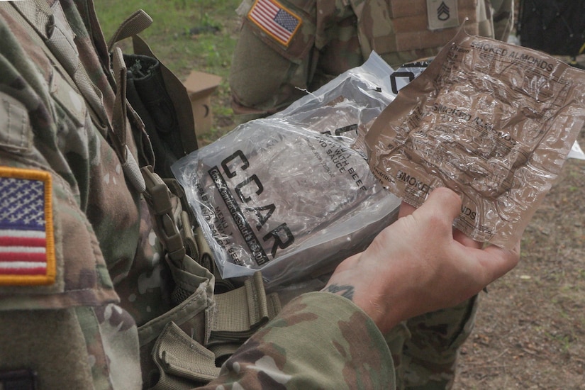 A person wearing a military camouflage uniform looks at two small plastic packages in their hands.