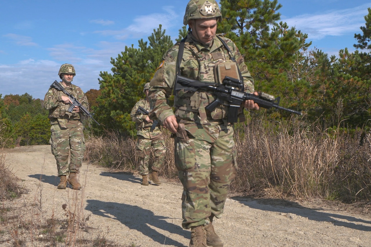 A soldier wearing a military camouflage uniform puts a small plastic package into a side pants pocket with one hand. Two other soldiers in similar attire walk behind him holding rifles.