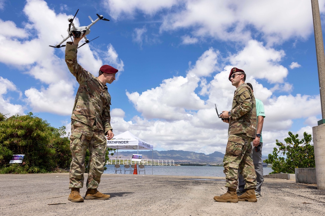 One soldier holds a remote control as another holds a drone toward a blue, cloudy sky with mountains in the distance.
