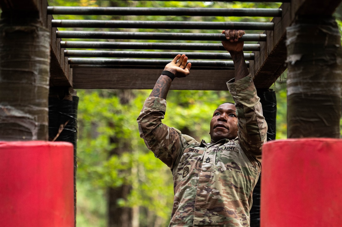 A soldier alternates hanging from bars while shifting one hand to the other outside on an obstacle course with a forest setting behind.