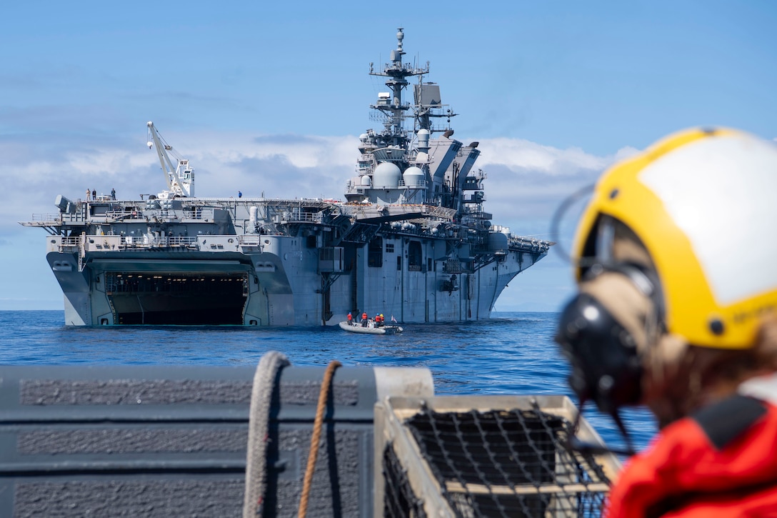 A small boat approaches a large ship as seen from another boat with a person wearing a yellow helmet to the right.