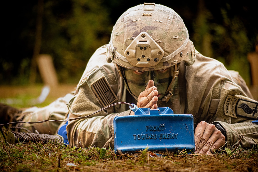 A helmeted and camouflaged soldier lays on grass and puts their hand on a blue mine with a wire running to the left while staring forward.