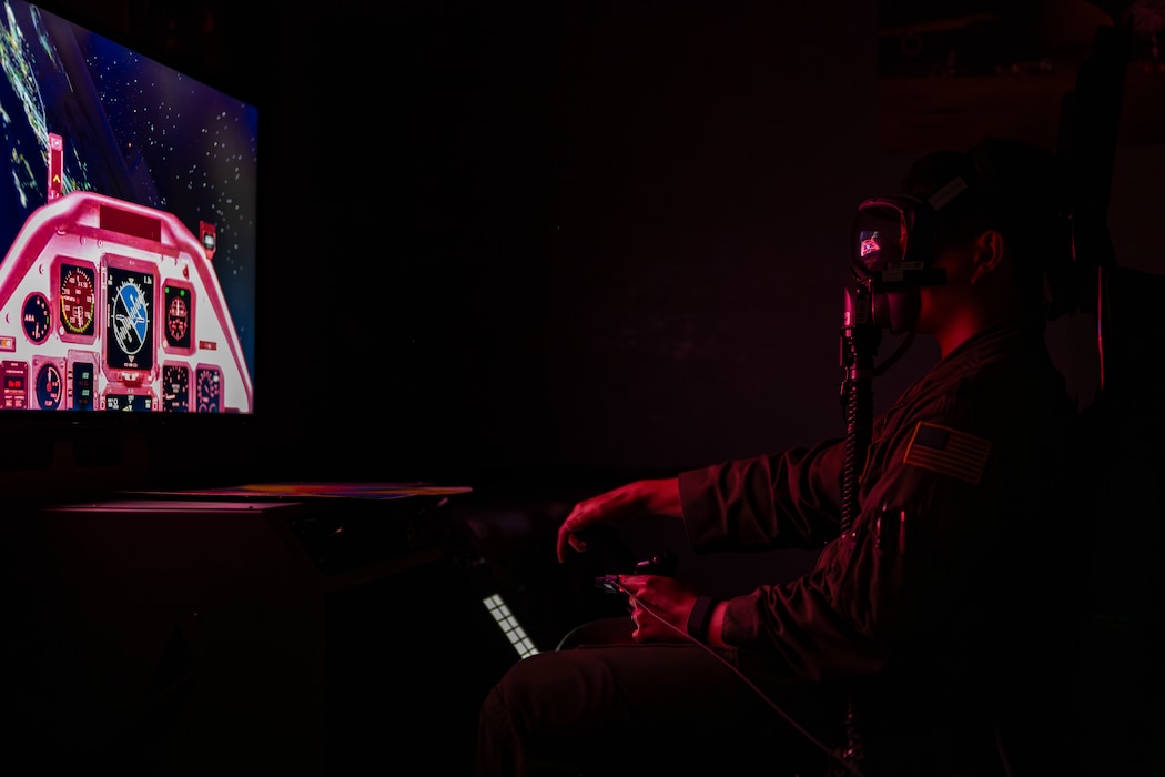 A screen on the left is visible with an airman on the right wearing a mask in a dark room illuminated by red light.