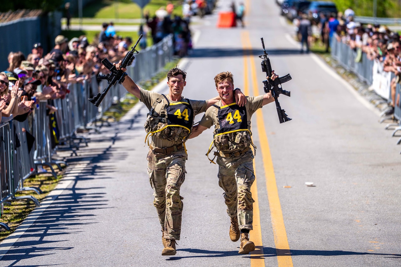 Two men in camouflage military uniforms run on a road with their arms around each other while holding rifles in the air. On either side of the road, long lines of spectators cheer them on.