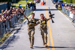 Two men in camouflage military uniforms run on a road with their arms around each other while holding rifles in the air. On either side of the road, long lines of spectators cheer them on.