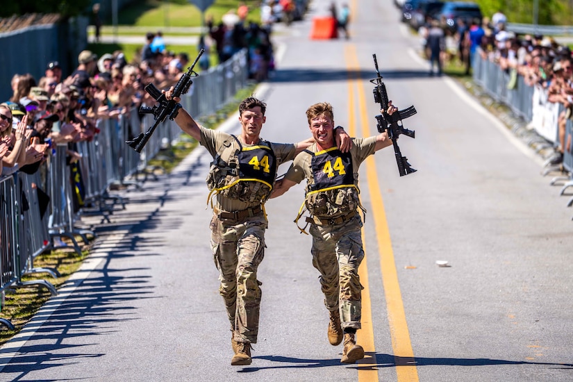 Two men in camouflage military uniforms run on a road with their arms around each other while holding rifles in the air. On either side of the road, long lines of spectators cheer them on.