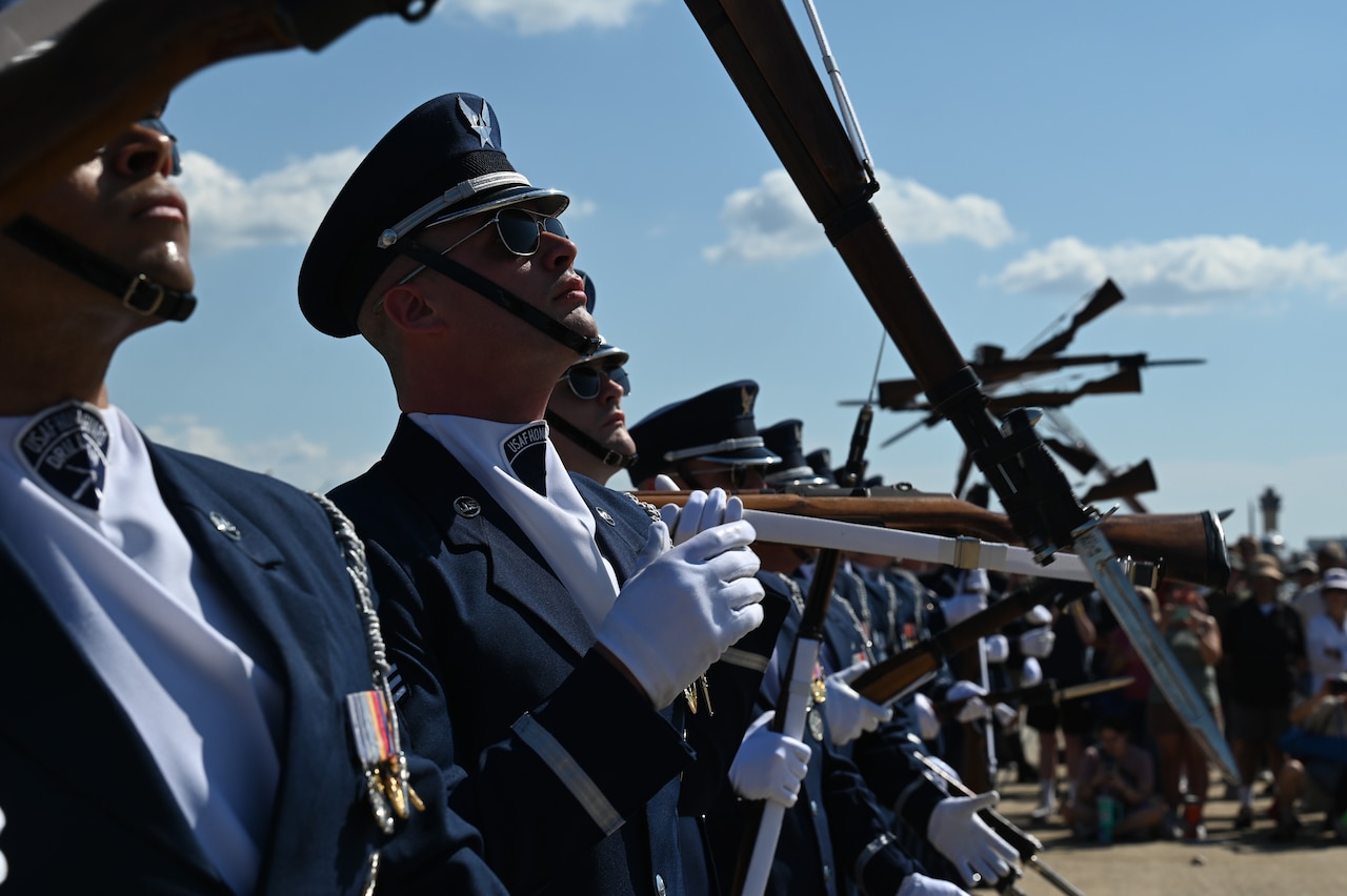 Airmen dressed in honor guard uniforms toss their rifles in the air while performing in a line formation on a tarmac.