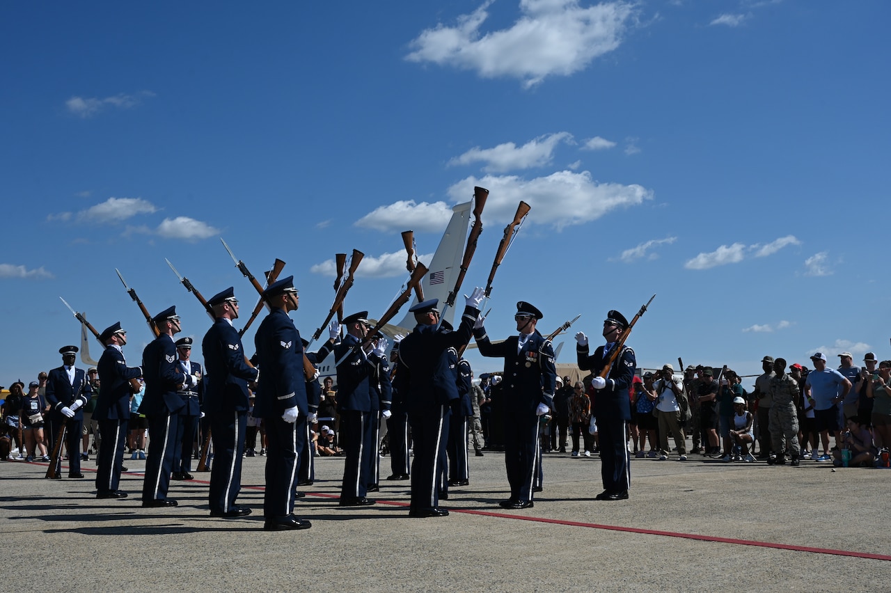 Airmen dressed in honor guard uniforms pass their rifles to each other during a performance on a tarmac. There are people in casual attire observing in the background.