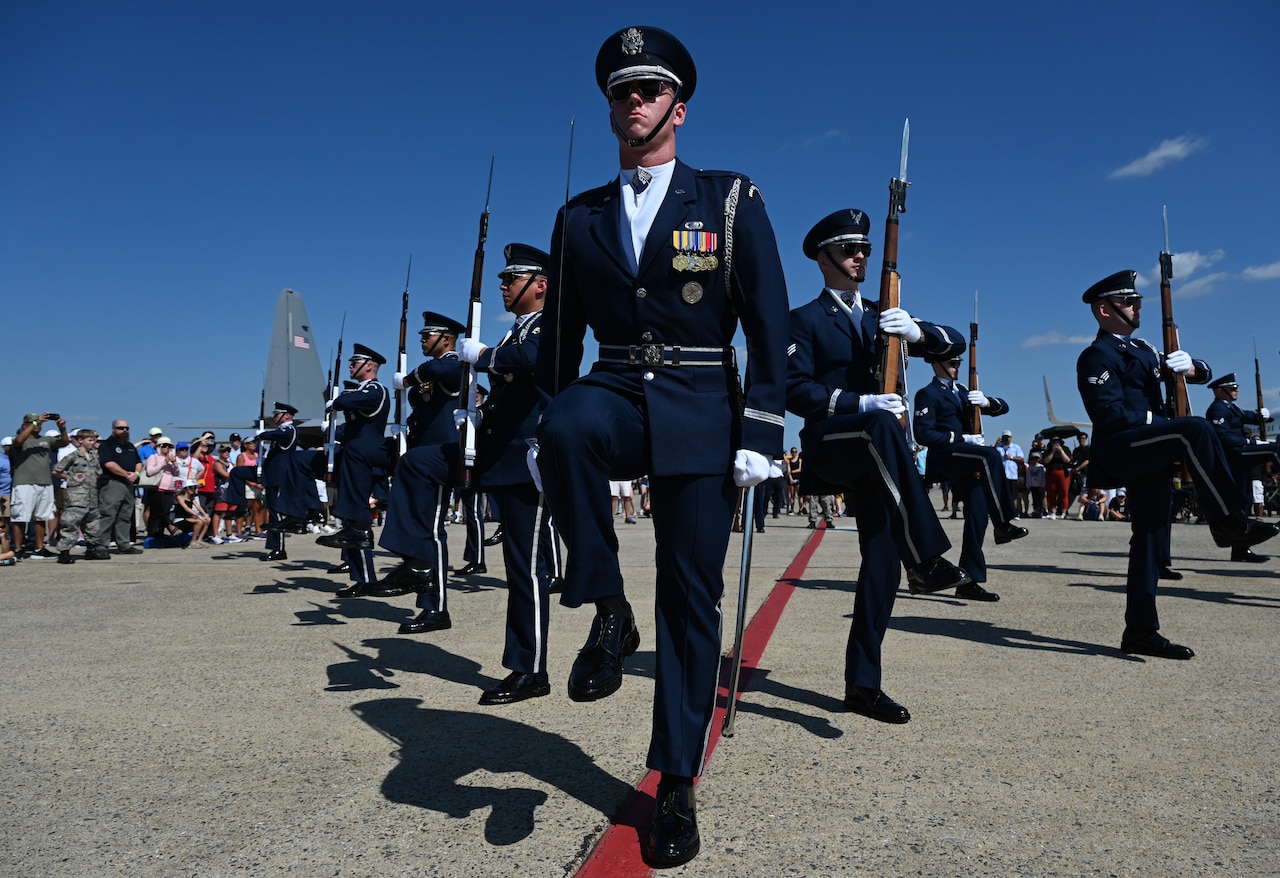 Airmen dressed in honor guard uniforms perform in a letter V formation. Several of the service members are holding rifles. There are people in casual attire observing in the background.