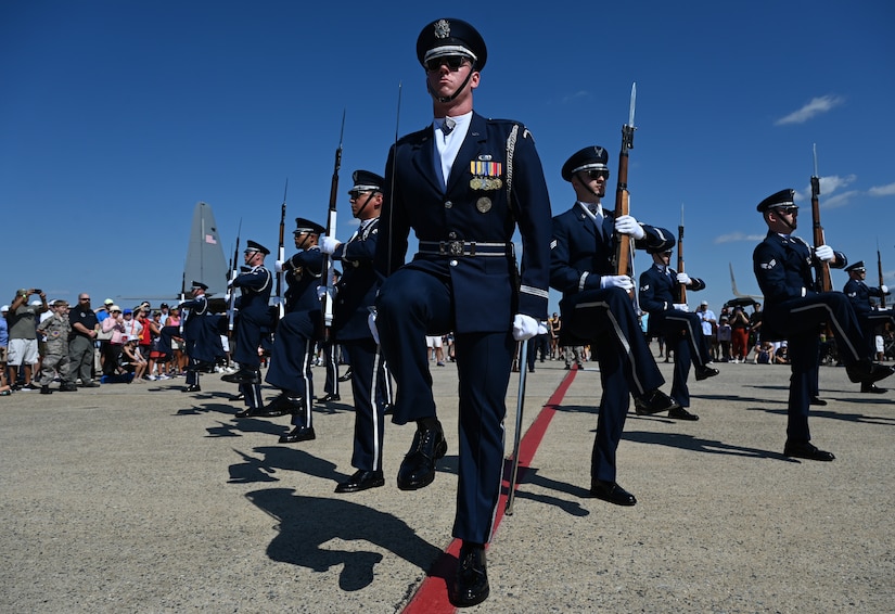 Airmen dressed in honor guard uniforms perform in a letter V formation. Several of the service members are holding rifles. There are people in casual attire observing in the background.