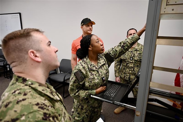Navy reservists participate in classroom instruction on radio communication systems as part of a NIWC Atlantic training program. An instructor is visible at the front of the room.