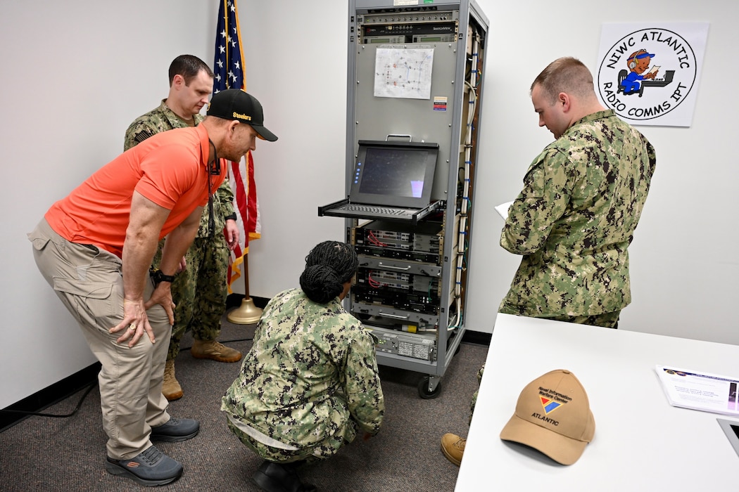 A group of U.S. Navy sailors and instructors work on electronic equipment in a lab setting. Wires and circuit boards are visible.