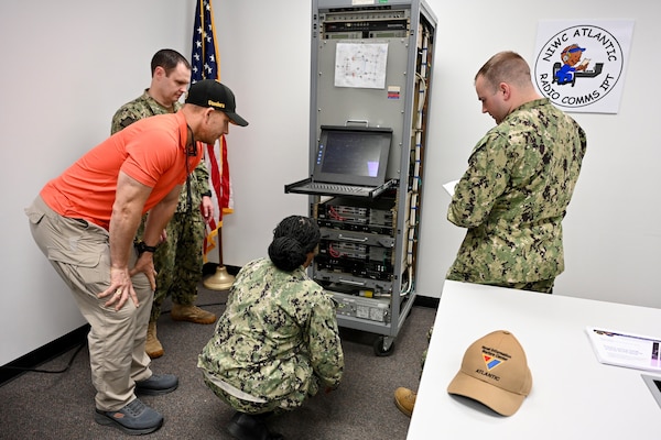 A group of U.S. Navy sailors and instructors work on electronic equipment in a lab setting. Wires and circuit boards are visible.