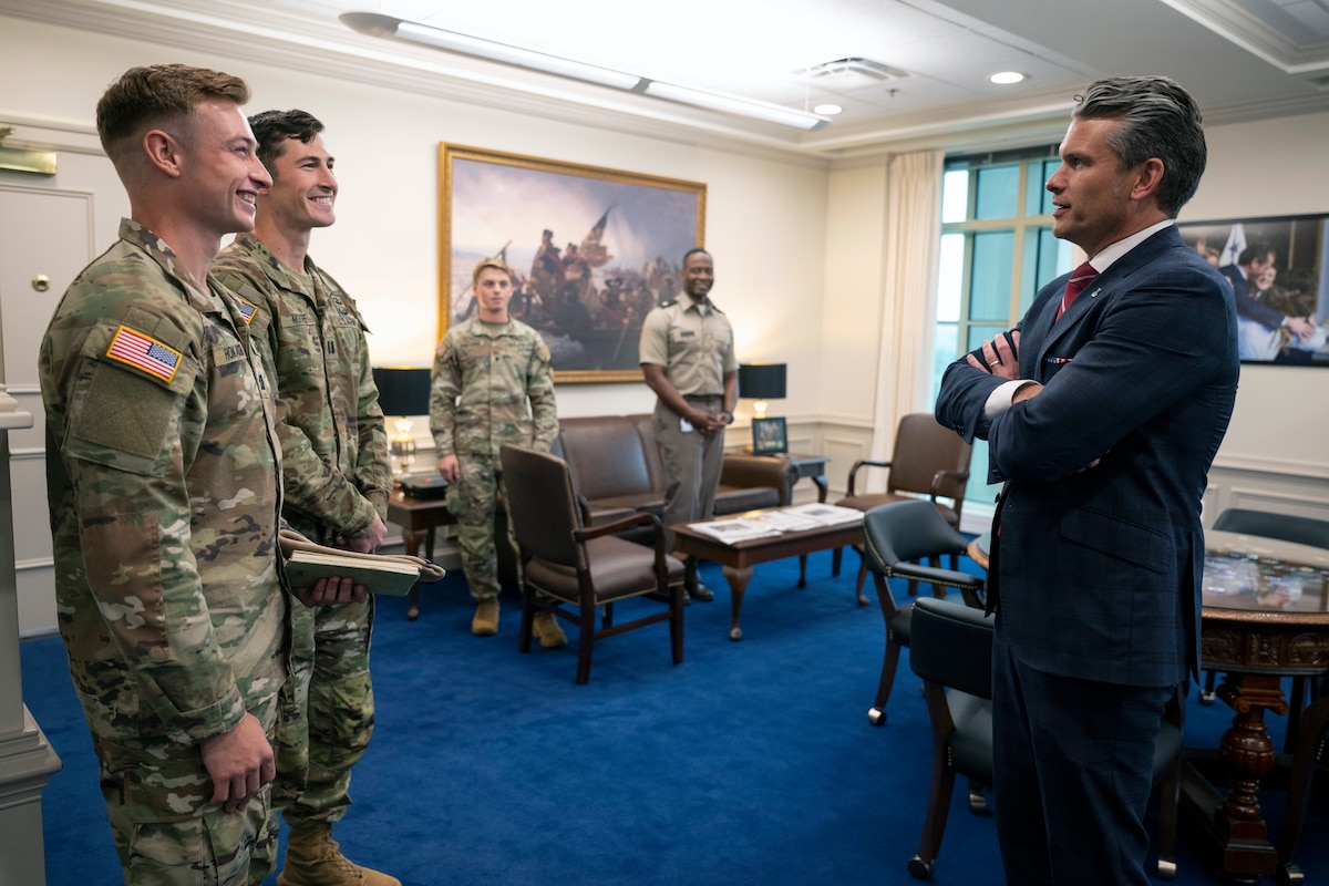 Secretary of War Pete Hegseth talks to two smiling soldiers while all stand in an office.