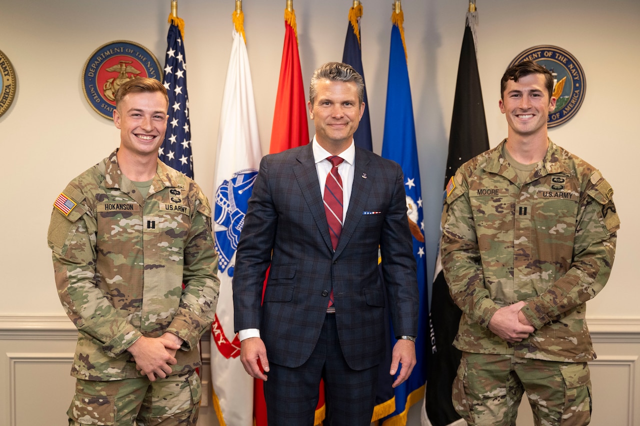 Secretary of War Pete Hegseth stands between two smiling soldiers in front of several flags in a room.