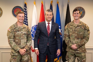Secretary of War Pete Hegseth stands between two smiling soldiers in front of several flags in a room.