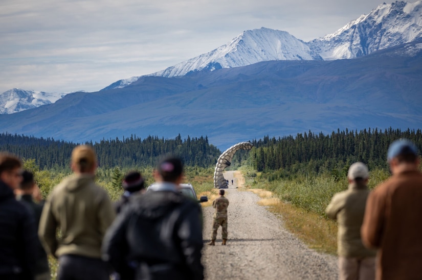 A group of soldiers and civilians watch supplies being air dropped onto a dirt road that runs through a wooded area.
