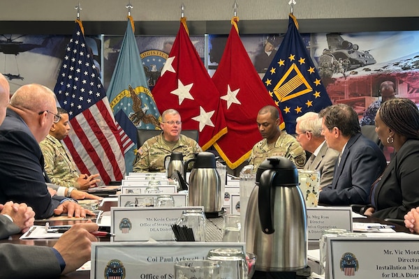 A large group of people sit around a conference table looking toward a man seated at the head of the table wearing a camouflage military uniform with military service flags behind him.