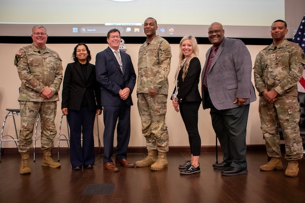 A group of seven people, three of which are wearing camouflage military uniforms, stand together on a stage posing for a group photo.