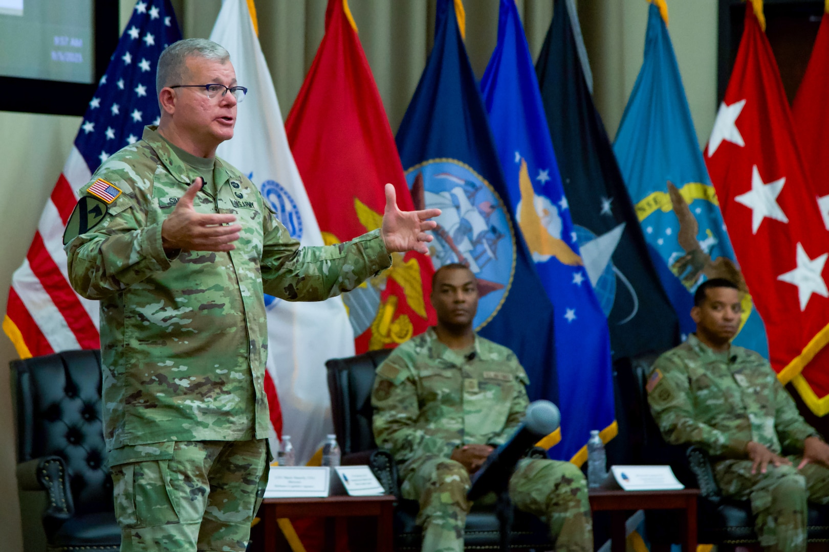 Three men wearing camouflage military uniforms are on a stage, two of them are seated with military service flags behind them and one is standing, speaking and gesturing to a crowd.