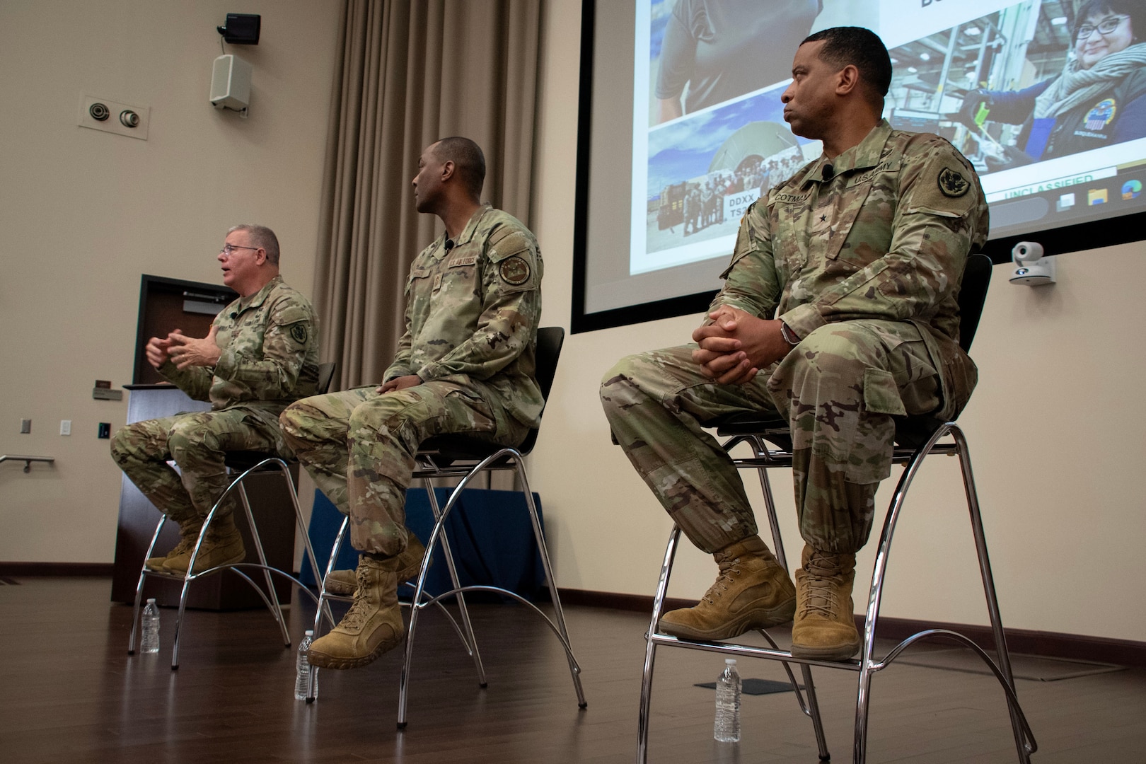 Three men wearing camouflage military uniforms sit on stools on a stage with a projection screen behind them.